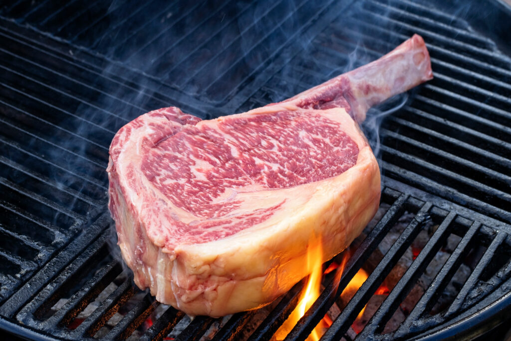 Raw dry aged Wagyu tomahawk steak offered as close-up on a charcoal grill with fire and smoke
