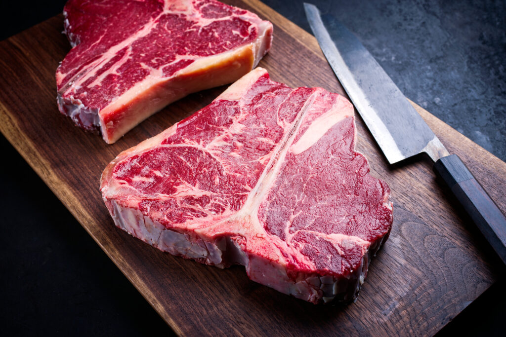 Two raw dry aged wagyu porterhouse beef steaks offered as close-up on modern design wooden board with a Japanese gyuto knife